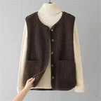 Dark brown sherpa fleece vest buttoned over a cream turtleneck jumper, hand folding back the collar to reveal suede lining