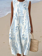 Close-up of a sleeveless midi dress in pale blue with a white leaf and branch print, carrying a straw tote