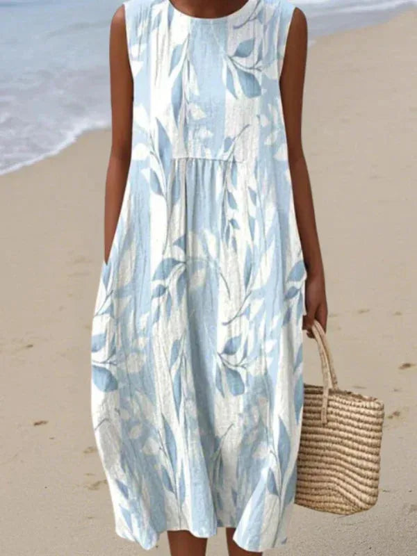 Close-up of a sleeveless midi dress in pale blue with a white leaf and branch print, carrying a straw tote