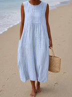Sleeveless midi dress in blue and white vertical stripes, paired with a woven straw bag on the beach