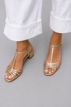 Close-up of gold strappy sandal with T-bar strap and low block heel, worn with white cuffed denim pants