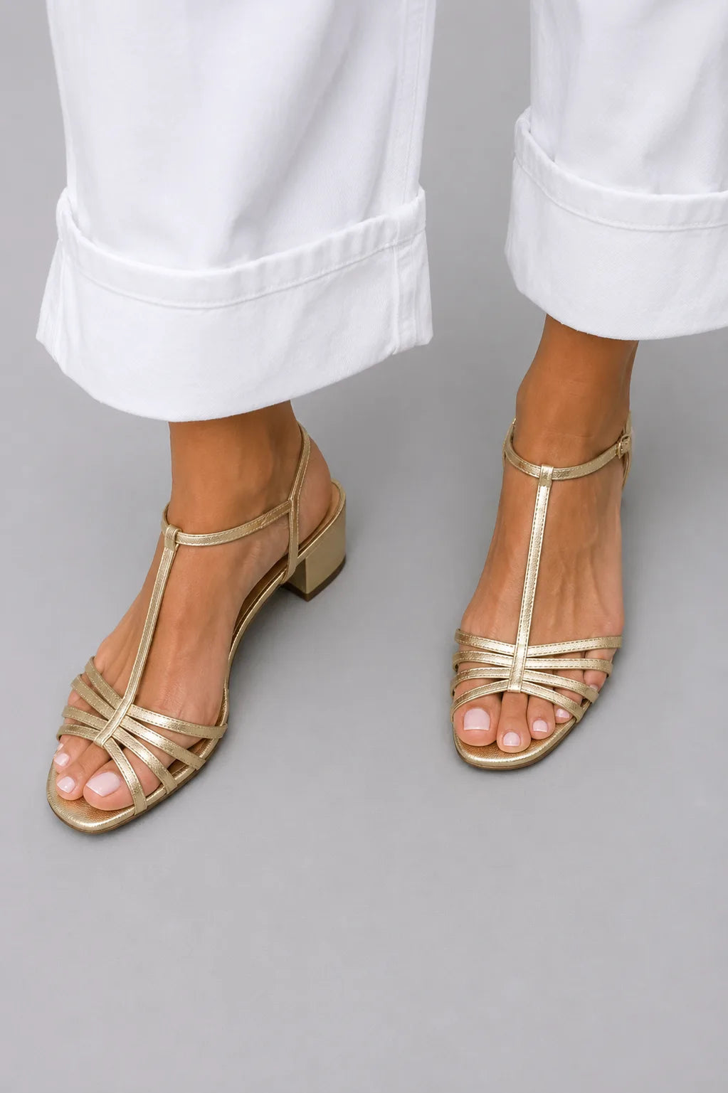 Close-up of gold strappy sandal with T-bar strap and low block heel, worn with white cuffed denim pants