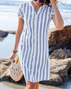 Model wears a blue and white vertical striped midi shirt dress with button placket, standing on a beach by the water