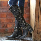 Close-up side view of black studded knee-high western boot showing dense stud embroidery pattern and stacked block heel against a brick wall