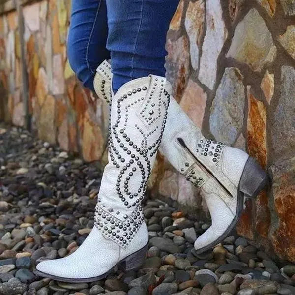Side view of white studded leather knee-high western boot with intricate stud pattern, pointed toe and side zip, against a stone wall