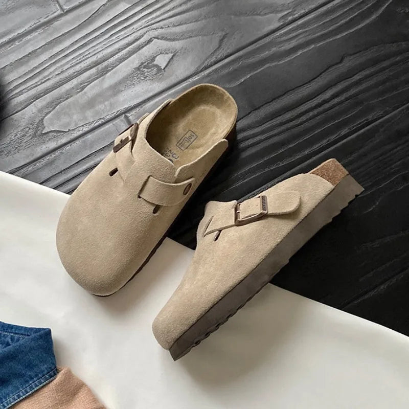 Beige suede clog slipper pair resting on a dark wood surface, showing buckle strap and stitched vamp