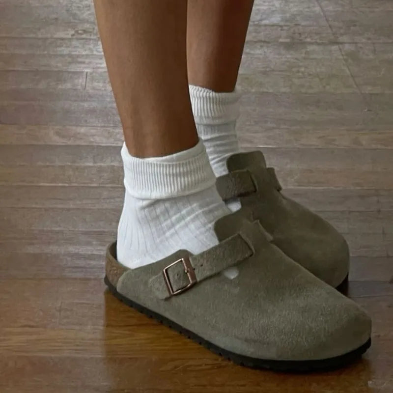 Model wearing olive green suede clogs with white crew socks, standing on a timber floor