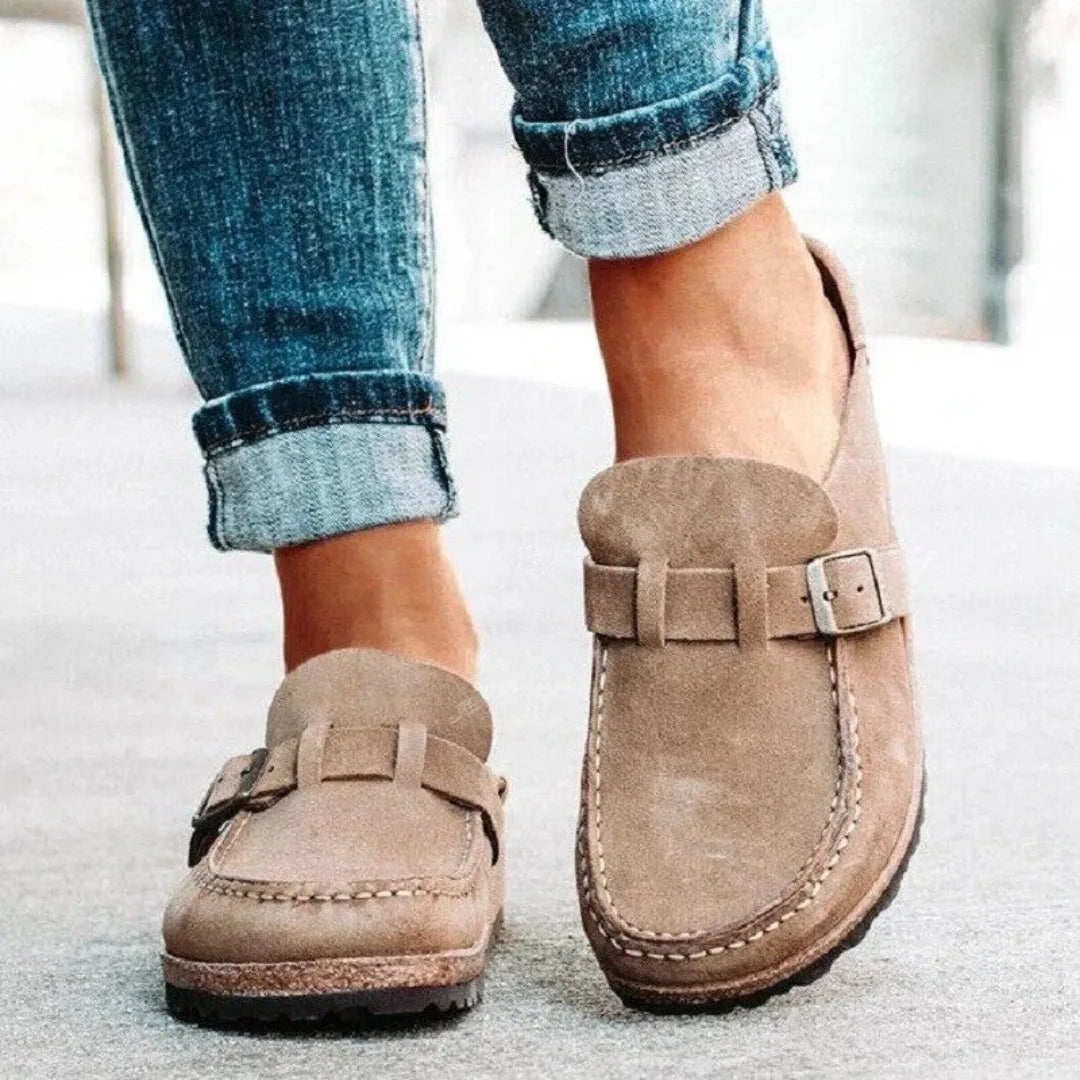 Side view of khaki suede slip-on clogs with buckle strap detail and visible topstitching, worn with cuffed jeans