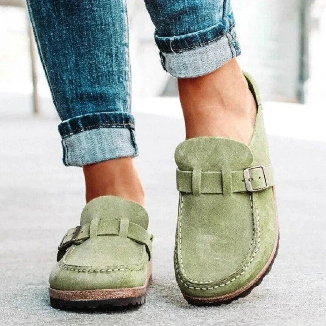Close-up of sage green suede backless loafers showing the metal buckle and contrast stitched toe seam
