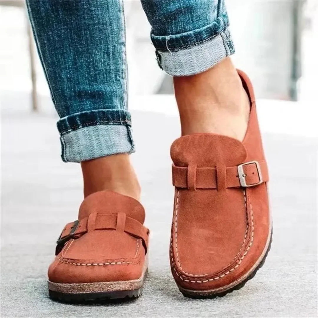 Rust red suede slip-on shoes with buckle trim shown from behind while walking in rolled-up denim