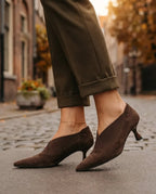 Side view of a woman's ankle wearing a brown suede kitten heel pump with V-shaped elastic gore, paired with cuffed olive trousers.