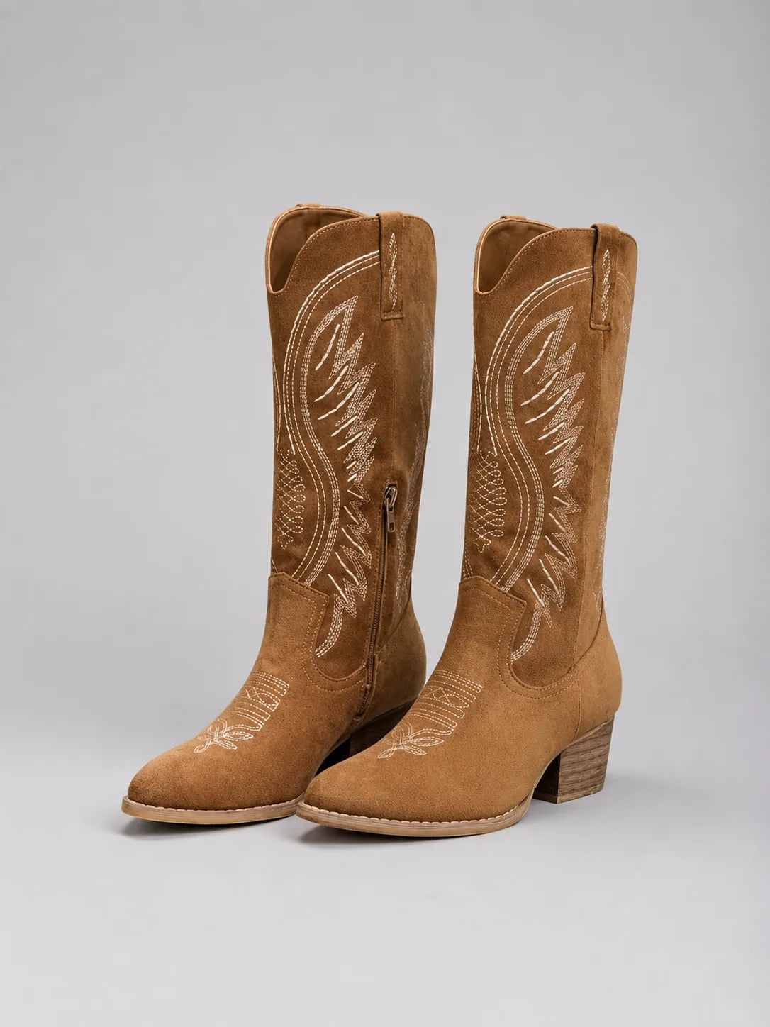 Pair of tan suede knee-high cowboy boots with cream western stitching, shown angled on a plain grey background