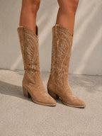 Close-up of tan suede cowboy boots on model's legs, showing the stacked block heel and embroidered shaft detail