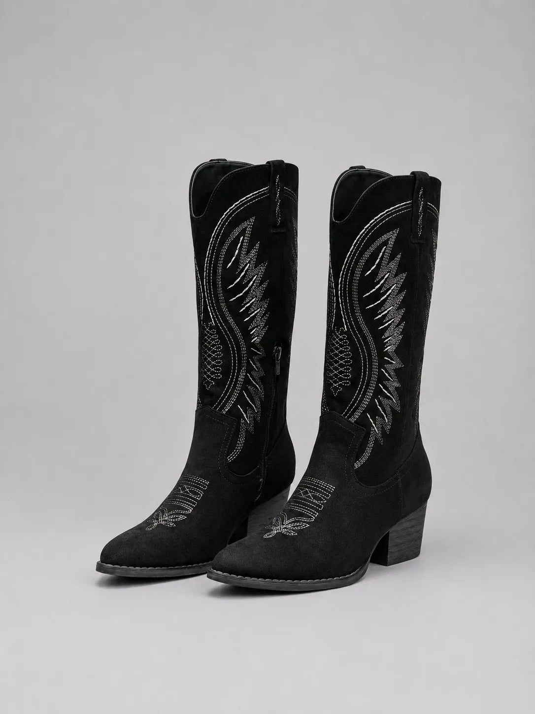 Pair of black suede knee-high cowboy boots with contrast stitching, shown angled on a plain grey background