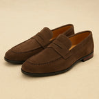 Pair of dark brown suede penny loafers shown at an angle, highlighting the apron stitching and tan leather lining