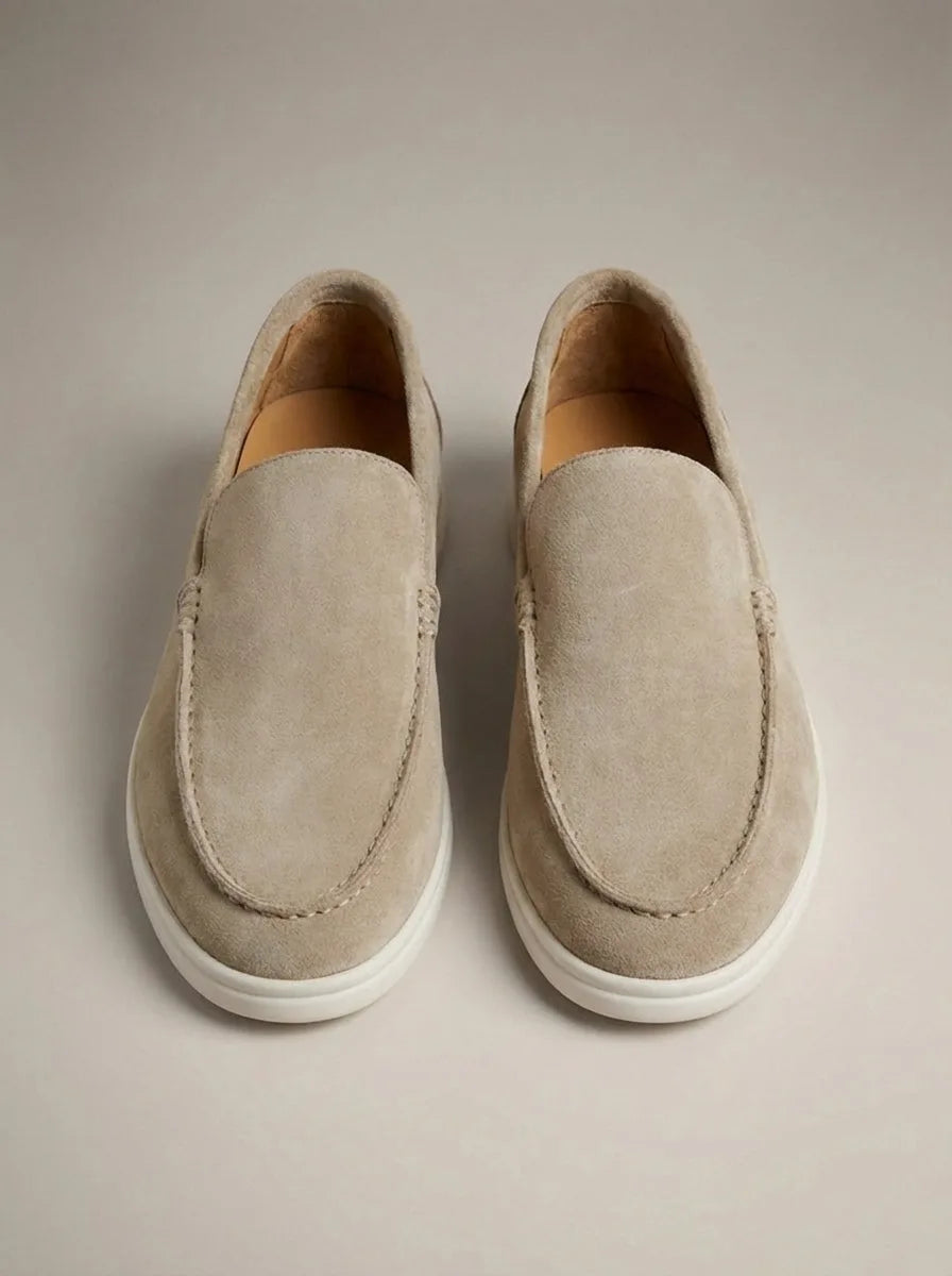 Top-down view of beige suede slip-on loafers with white outsole and tan leather lining on a neutral backdrop