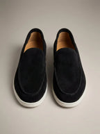 Pair of black suede slip-on loafers shown from above, featuring a moc-toe seam and contrasting white sole