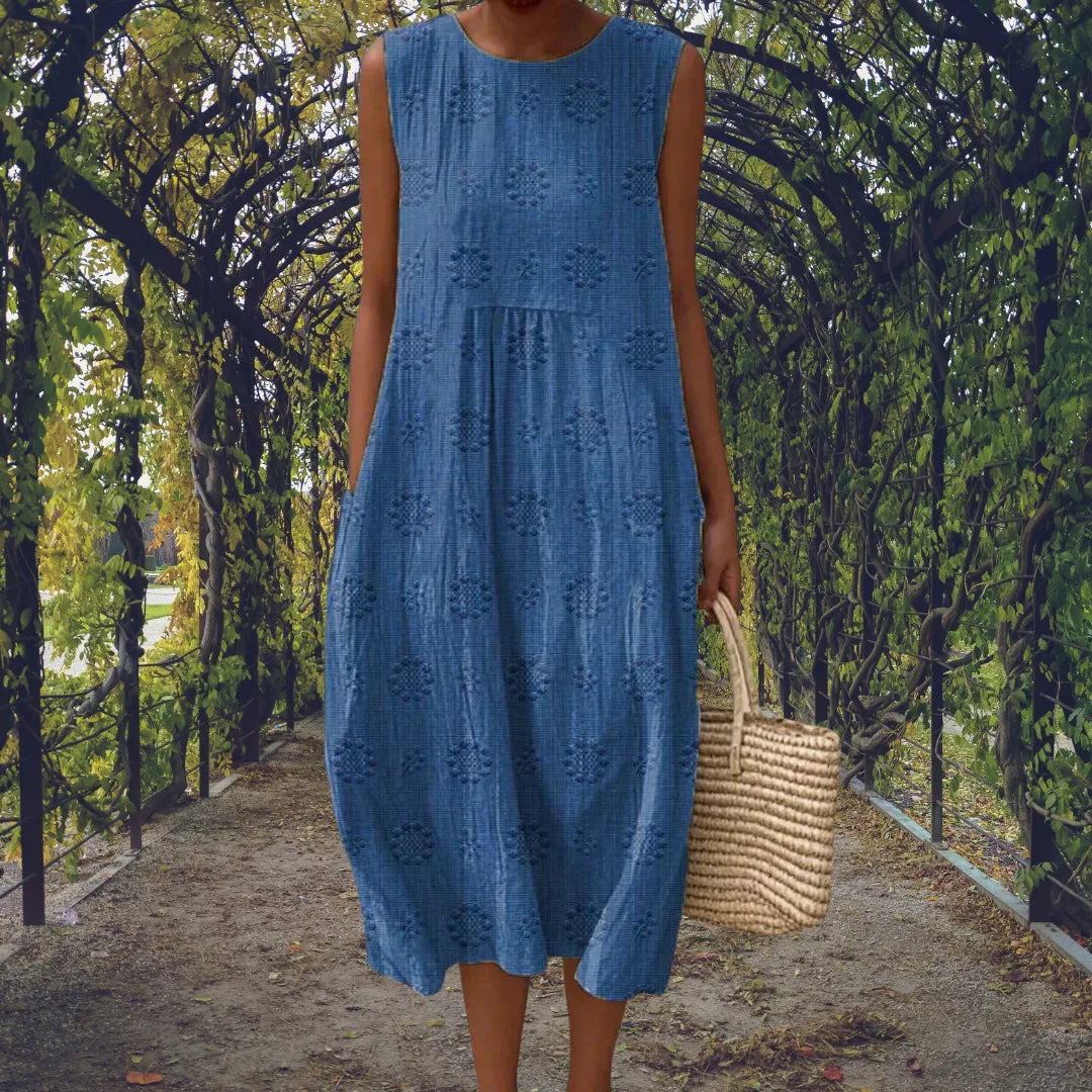 Blue sleeveless midi dress with textured tonal dot embroidery, styled with a woven straw tote bag.