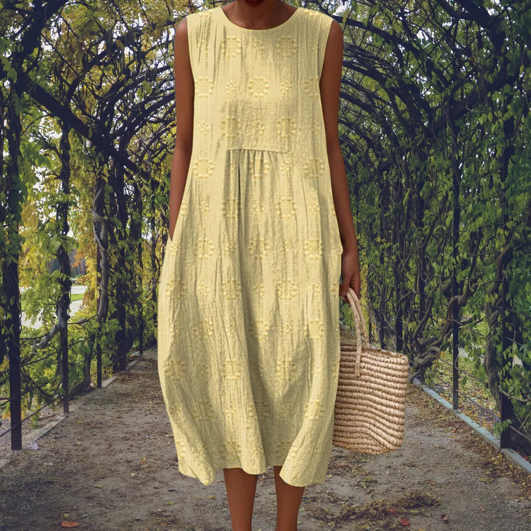 Pale yellow sleeveless midi dress with a gathered front panel, shown walking along a tree-lined path.