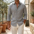 Man wearing a heathered grey long-sleeve shirt, unbuttoned at the collar, paired with cream trousers in a sunlit courtyard