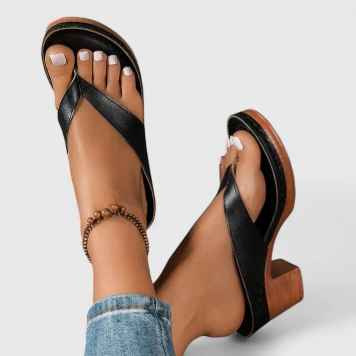 Black toe-post sandals with wooden block heel, seen from above on crossed feet wearing an anklet