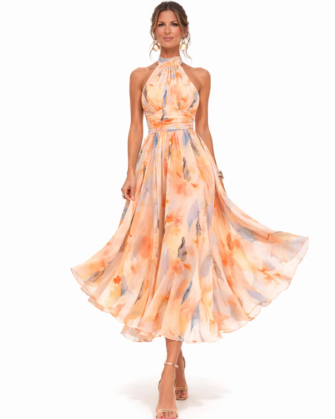 Front view of a model walking in a flowing peach floral print halter dress with gold sandals