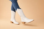 White faux leather cowboy boots worn with jeans, mid-stride showing pointed toe and stacked heel