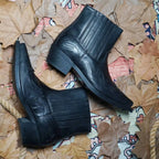 Overhead flat-lay of a pair of black leather western ankle boots resting on autumn leaves and a woven rug