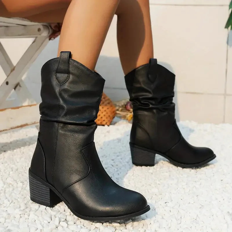 Black slouch western boots with ruched leather upper and stacked heel, worn outdoors on a textured rug
