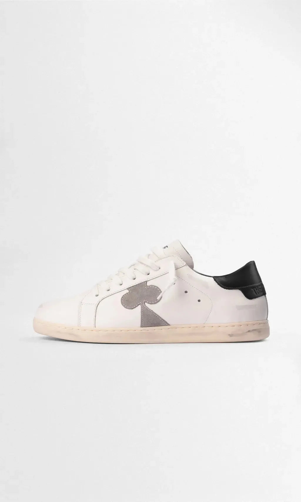 Side profile of a white leather sneaker with a grey suede clover emblem and black leather heel tab.