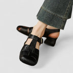 Close-up front view of black woven leather T-strap sandal with brass buckle and block heel, worn with jeans
