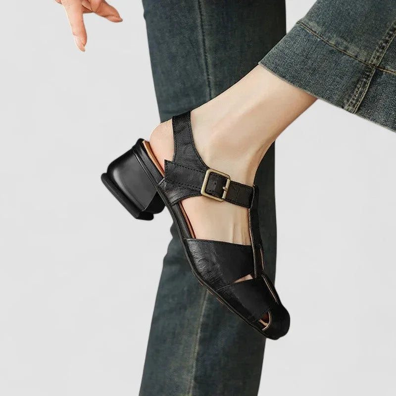 Rear view of burgundy leather sandals worn on both feet, showing block heels and ankle straps with black trousers