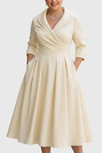 Front view of a shawl-collar wrap-style midi dress in pale beige satin with a pleated full skirt and side pockets, worn with a diamond necklace.