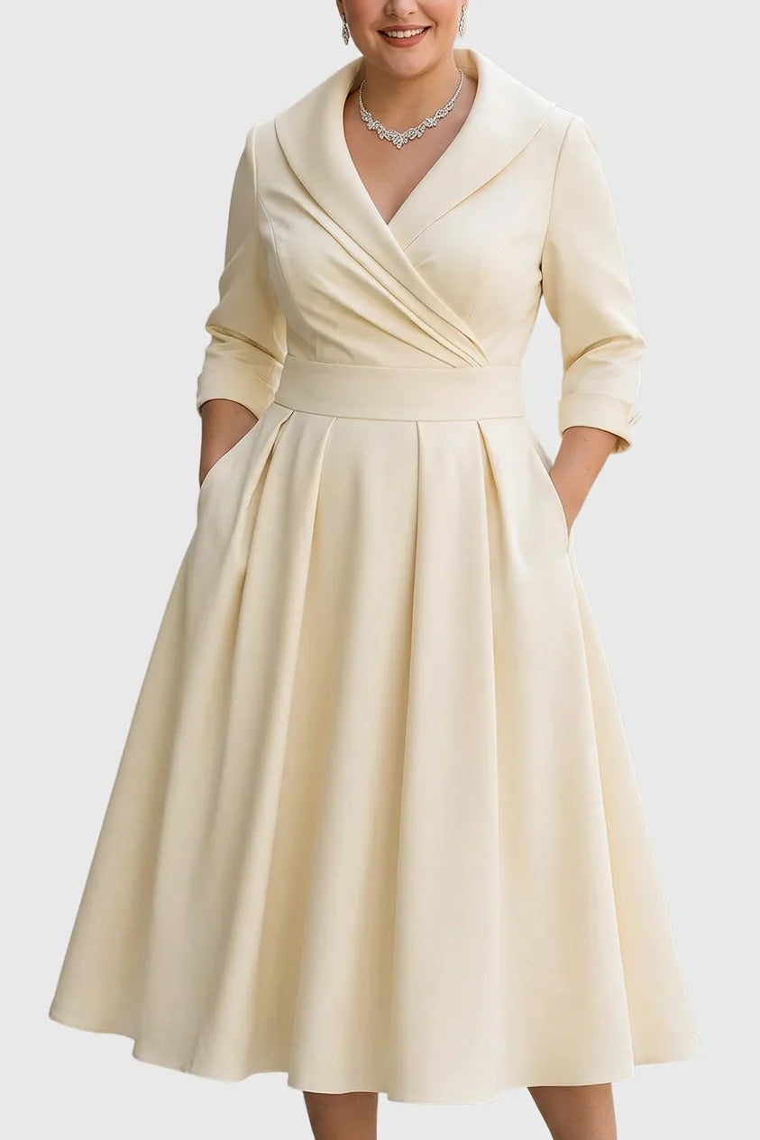 Front view of a shawl-collar wrap-style midi dress in pale beige satin with a pleated full skirt and side pockets, worn with a diamond necklace.