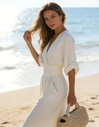 White wrap jumpsuit with gathered waistband styled casually on a beach with a woven straw bag